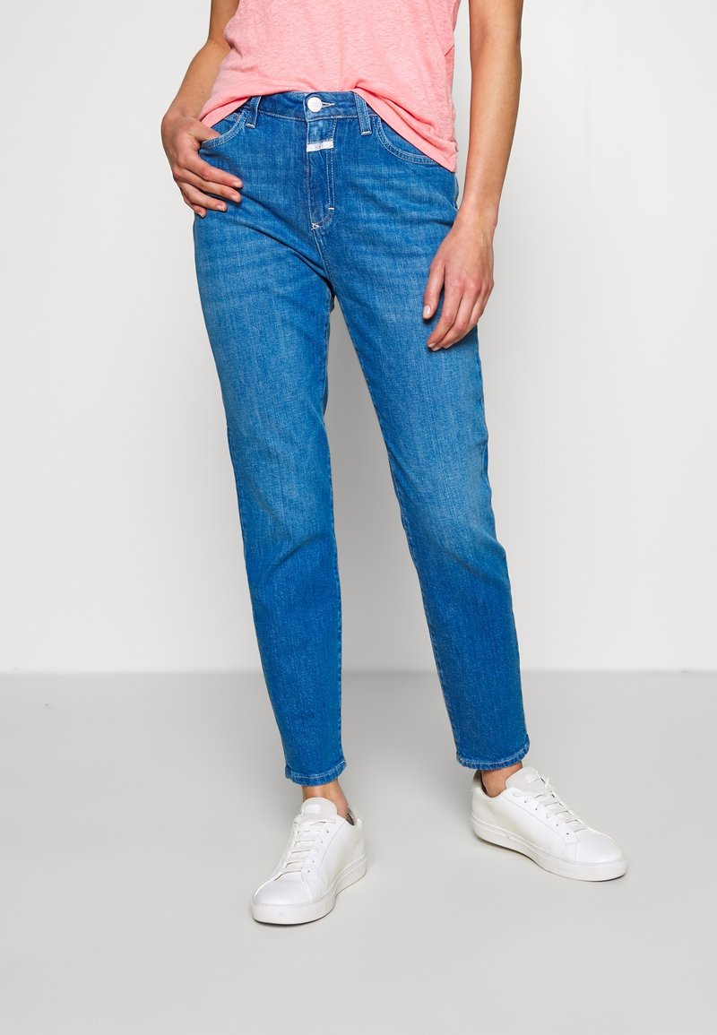 Femme portant un jean droit bleu moyen, un t-shirt rose rentré et des baskets blanches, debout devant un fond clair uni.