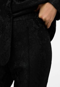Tissu en dentelle noire avec des motifs floraux, surface texturée, et une coupe ajustée avec un détail de poche latérale. Fermeture à boutons visible.
