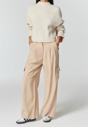 Personne portant un pull en maille crème, un pantalon cargo beige à jambes larges avec des poches, et des baskets noir et blanc, debout les mains dans les poches.