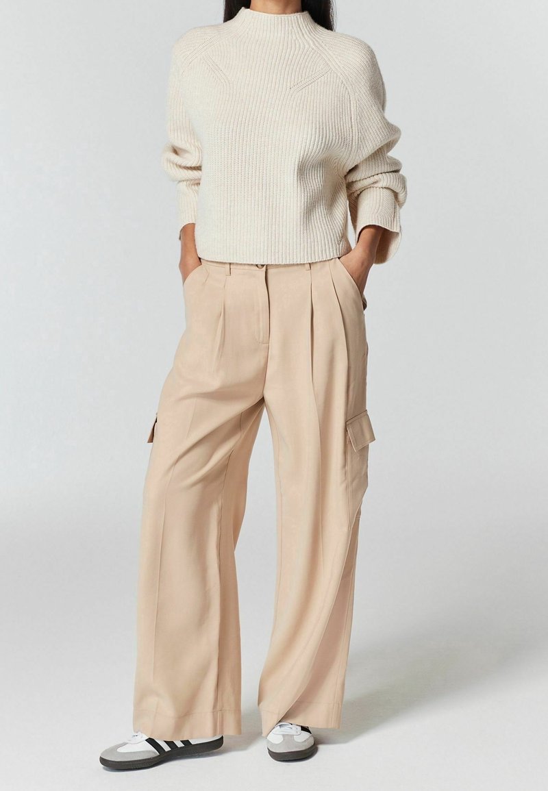 Personne portant un pull en maille crème, un pantalon cargo beige à jambes larges avec des poches, et des baskets noir et blanc, debout les mains dans les poches.