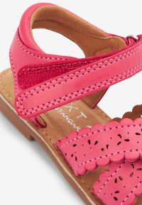 Rosa læder sandal med bølgede stropper, perforerede detaljer og justerbar Velcro-lukning. Brun tekstureret sål.