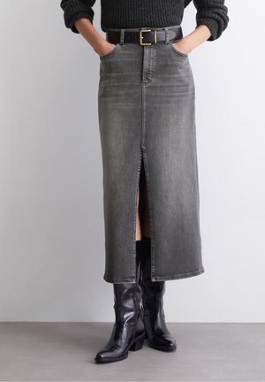 Kvinde iført en sort sweater, høj taljet falmet grå denimnederdel med frontslids, sort bælte og sorte læderstøvler til midt på læggen. Hænderne i lommerne.