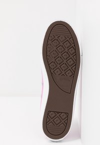 Baskets en toile rose avec une semelle en caoutchouc blanche. La semelle présente un motif brun texturé avec un logo au talon. Des œillets métalliques sont visibles.