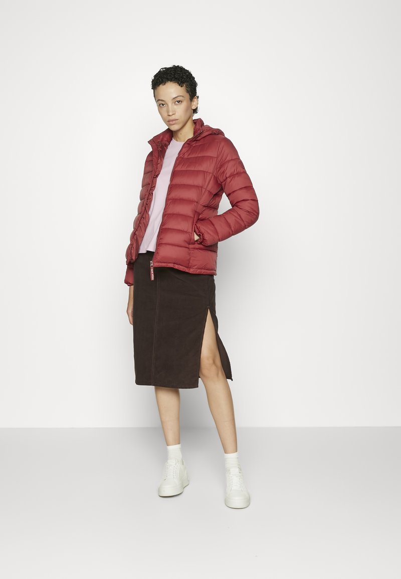 Chaqueta de plumas roja con capucha, cierre de cremallera y textura acolchada, combinada con una blusa rosa claro y una falda de ante marrón con una abertura lateral.