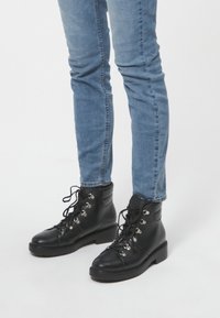 Stivaletti neri in pelle con punta tonde e occhielli argentati, allacciati sul davanti. Indossati con jeans in denim blu aderenti.