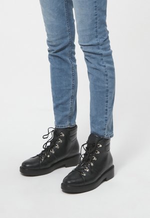 Bottines en cuir noir avec des orteils ronds et des œillets en argent, lacées à l'avant. Portées avec un jean en denim bleu slim-fit.