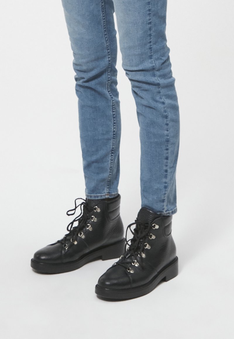 Stivaletti neri in pelle con punta tonde e occhielli argentati, allacciati sul davanti. Indossati con jeans in denim blu aderenti.