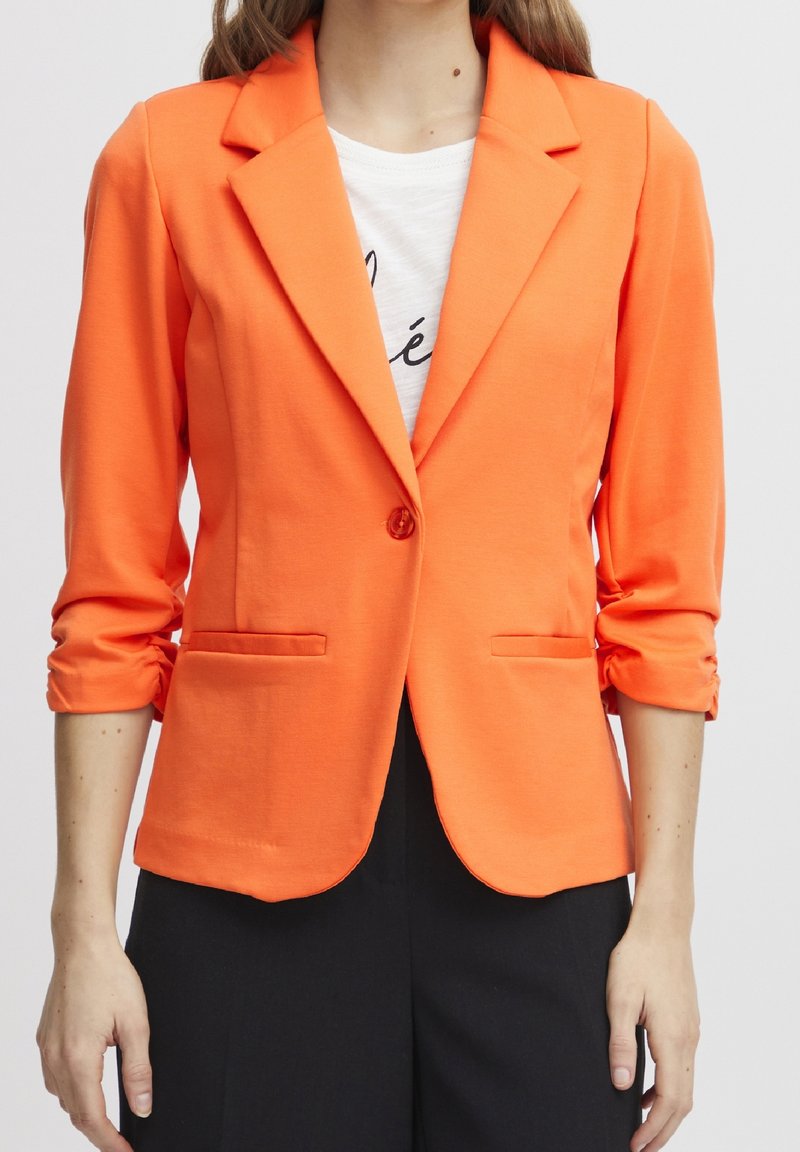 Femme portant un blazer orange vif avec un seul bouton sur une chemise blanche et un pantalon noir, debout devant un fond uni.