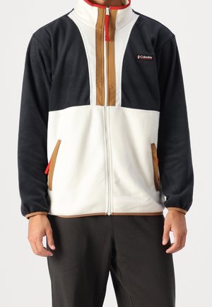 Personne portant une veste polaire de la marque Columbia noire, blanche et marron avec une fermeture éclair à l'avant et deux poches bordées de marron.