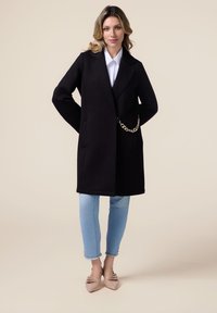 Cappotto in misto lana nero con design doppiopetto, tasche laterali e dettaglio a catena. Abbinato a jeans blu chiaro e scarpe beige a punta.