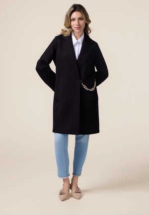 Cappotto in misto lana nero con design doppiopetto, tasche laterali e dettaglio a catena. Abbinato a jeans blu chiaro e scarpe beige a punta.