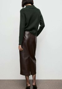 Mujer vestida con un suéter acanalado verde oscuro, una falda midi de cuero marrón oscuro con una abertura en la espalda y zapatos de tacón negros, de pie contra una pared blanca.