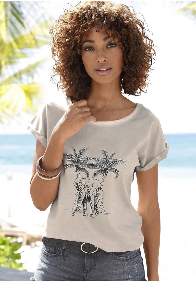 Chemise beige à manches courtes avec un graphisme d'éléphant et de palmiers. Portée avec un short en denim foncé et une ceinture tressée.