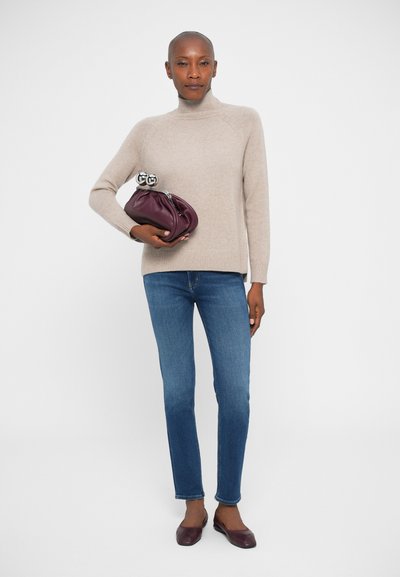 Vrouw in een beige coltrui, blauwe jeans en bordeauxrode ballerina's, die een bijpassende bordeauxrode handtas vasthoudt tegen een witte achtergrond.