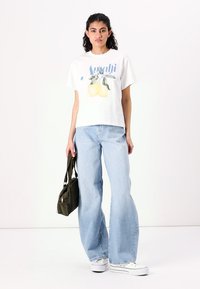 Hvid t-shirt med blå grafisk tekst og citronillustration, parret med lyseblå bredbenede jeans og hvide sneakers; holder en grøn taske.