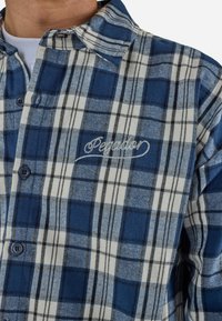 Gros plan d'une chemise à carreaux bleue et blanche avec un col et un texte brodé sur la poitrine, portée sur un sous-vêtement blanc.