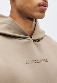 Κοντινό πλάνο ενός ατόμου που φοράει μπεζ φούτερ με κουκούλα με το ανάγλυφο γράψιμο "J.LINDEBERG" στο στήθος, εμφανίζοντας το κάτω μέρος του προσώπου και τον λαιμό.