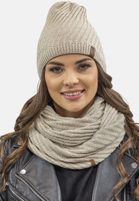 Beige ribbet strik beanie med en afslappet design og et lille mærke; matchende chunky strikket infinity tørklæde; blød tekstur; båret med en sort læderjakke.