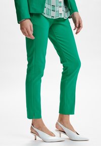 Zielone spodnie o dopasowanym kroju z bocznymi rozcięciami, zestawione z białymi czółenkami w szpic, ukazujące gładką fakturę materiału.
