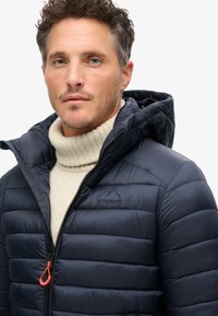 Mørkeblå puffy jakke med en quiltet tekstur, lynlås lukning og hætte. Har et broderet logo på brystet og en lys lynlås træk.