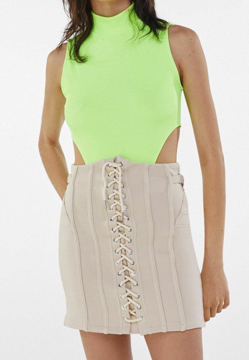 Bodysuit côtelé vert néon avec un col montant et des emmanchures découpées, associé à une mini-jupe beige avec des coutures verticales et un devant à lacets.