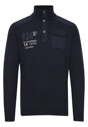 Mørkeblå strikpullover med høj krave, trykknapper, skulderdetaljer og en brystlomme. Har broderet tekst nær lommen.