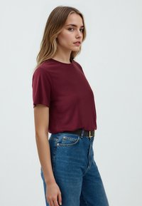 T-shirt corto di colore marrone con scollo rotondo, indossato con jeans blu a vita alta. Tessuto morbido e vestibilità comoda, senza piante o dettagli.