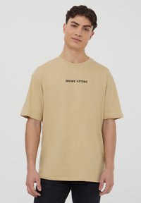 T-shirt beige con maniche corte, dotato di scollo a girocollo e con la scritta "WEST COAST" in un carattere nero a contrasto sul davanti.