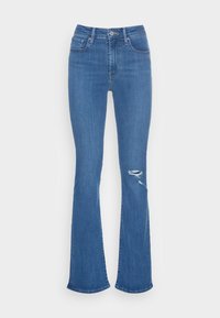 Jean en denim bleu taille haute avec légère évasement et déchirure usée sur la cuisse droite, doté de poches avant et arrière ainsi que d'une fermeture à boutons.