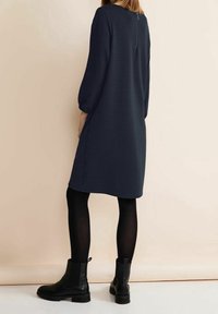 Kvinna iklädd en knälång, långärmad marinblå klänning, svarta strumpbyxor och svarta ankelstövlar, stående mot en beige bakgrund.