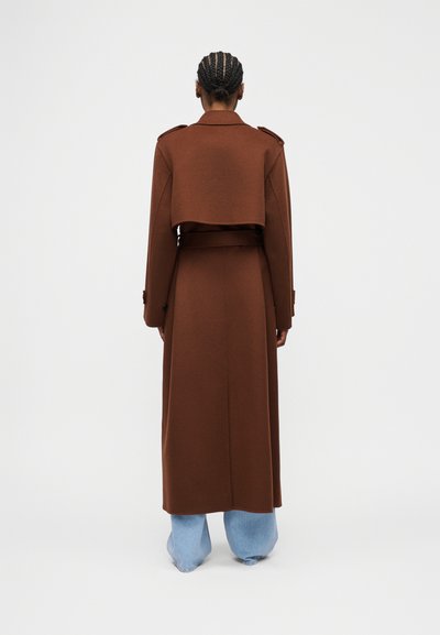 Άτομο με πλεξούδες φορά ένα μακρύ καφέ παλτό με κοντή κάπα στο πίσω μέρος, συνδυασμένο με ανοιχτόχρωμο μπλε τζιν με φαρδιά πόδια, στέκεται μπροστά από ένα απλό φόντο.