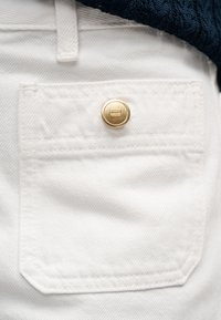 Pantalon en coton blanc avec une poche arrière ornée d'un bouton doré et un tissu subtilement texturé. La poche présente un bord droit et des détails cousus.
