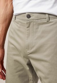Chinos in cotone verde chiaro con fronte piatto, chiusura con un solo bottone e tasca laterale. Tessuto liscio con dettagli di cuciture visibili.