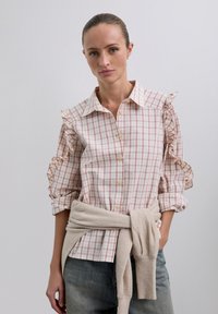Camisa de cuadros blanca y rosa con mangas rizadas y botones en el frente. Suéter beige claro atado a la cintura, combinado con jeans azules.