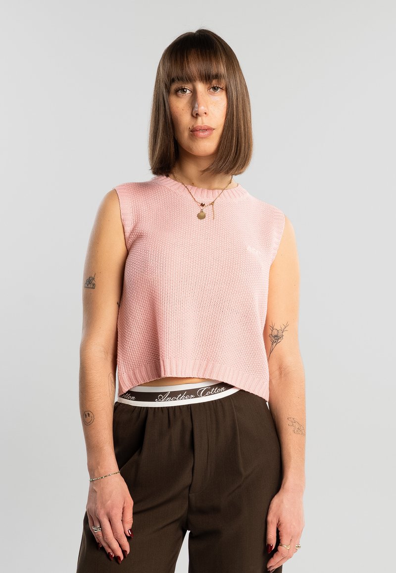 Femme aux cheveux bruns raides porte un haut en tricot rose sans manches, un pantalon sombre taille haute, une ceinture "Another Cotton" visible, et plusieurs tatouages sur les bras.
