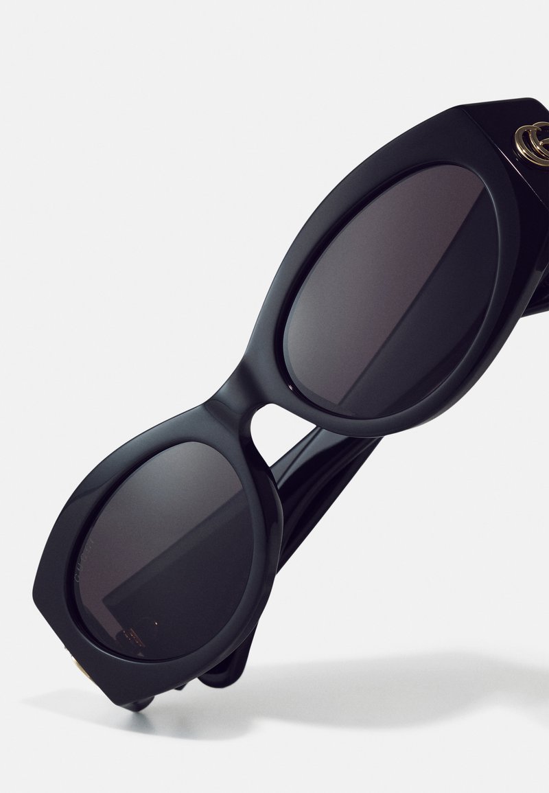 Schwarze übergroße Sonnenbrille mit einer glatten, matten Oberfläche, ausgestattet mit grauen Gläsern, einer ausgeprägten geometrischen Form und goldenen Hardware-Details.