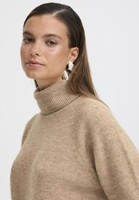 Maglione beige a collo alto con una texture morbida, colletto a costine visibile e maniche raglan. Orecchini geometrici argentati aggiungono un dettaglio contemporaneo.