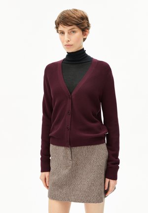 Vrouw met kort bruin haar, gekleed in een donkerpaarse cardigan met knopen, een zwarte turtleneck en een gestructureerde grijze rok, staande tegen een witte achtergrond.