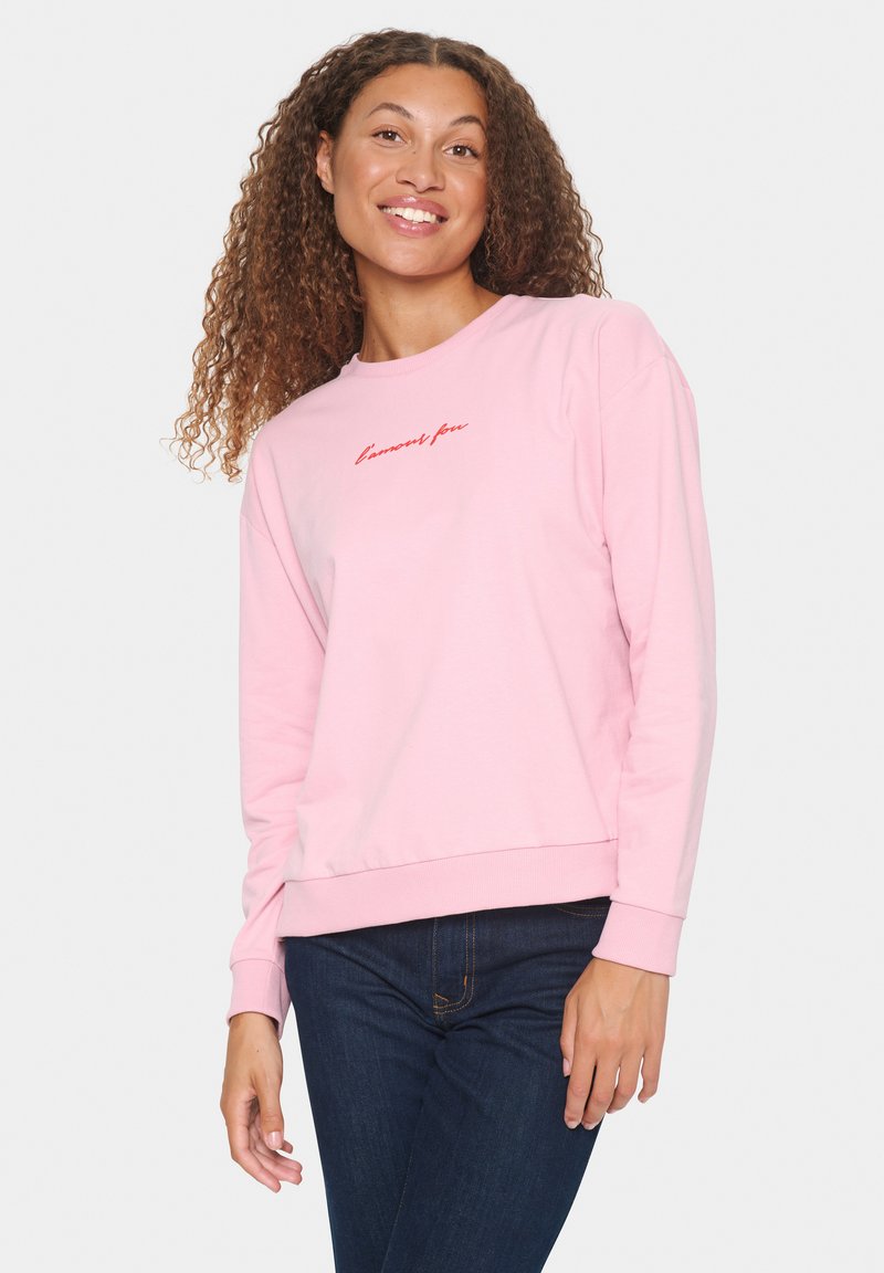 Sudadera rosa de manga larga, con cuello redondo, y texto rojo "l'amo(n)feu" en el pecho. Tejido suave y corte holgado. Combinada con vaqueros oscuros.