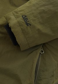 Olivegrüner Anorak aus strukturiertem Stoff, mit Reißverschlusstasche und schwarzen elastischen Bündchen. Auf dem Ärmel befindet sich ein kleines gesticktes Logo.