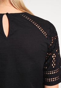 Svart blus med genomskinliga spetsdetaljer på korta ärmar och nyckelhålsringning. Mjukt tyg med lätt glans och subtil textur.