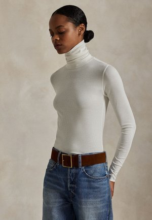 Kvinde med mørkt hår sat tilbage, iført en hvid højhalset trøje og blå jeans med et bredt brunt bælte, stående foran en neutral baggrund.