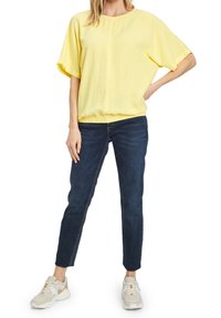 Żółta bluzka z krótkim rękawem, okrągłym dekoltem i elastycznym dołem, w połączeniu z ciemnoniebieskimi dżinsami i białymi sneakersami.