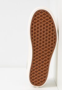 Biała podeszwa sneakerów z brązowym gumowym bieżnikiem z wzorem w diamenty i heksagony, ustawiona pionowo na białej powierzchni.