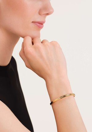 Pulsera de metal dorado con un diseño liso y redondeado, que presenta sutiles acentos decorativos y un acabado pulido.