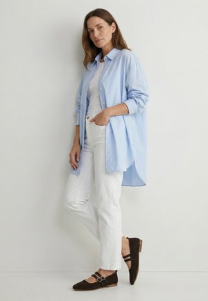 Frau trägt ein hellblaues, übergroßes Hemd über einem weißen Top und weißen Hosen, steht mit einer Hand in der Tasche an einer schlichten Wand.