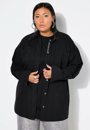 Kvinde med mørkt hår bærer store ringformede øreringe, en sort oversized skjorte med knapper og sølvglitrende bukser, stående mod en hvid baggrund.