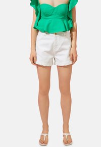 Top peplum verde con mangas cortas con volantes, combinado con pantalones cortos de mezclilla blanca con dobladillos deshilachados. Chanclas blancas completan el look.