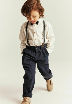Young child wearing white shirt, black bow tie, suspenders, dark pants, and tan shoes, looking down with hands in pockets while stepping forward.