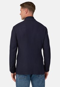 Mörkblå kavaj med en strukturerad design, snittad slag, enkel ryggspringa och subtil herringbone-textur. Har knappade manschetter.